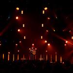 Trivium lead-singer Matt Heafy on a dimly-lit stage playing an acoustic guitar in front of a large crowd.